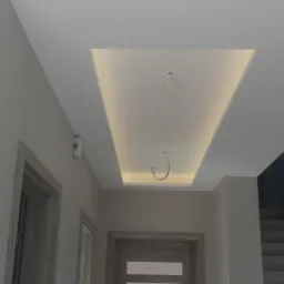 Nowoczesny sufit podwieszany z oświetleniem LED w kształcie prostokąta, widoczne przewody elektryczne przed ostatecznym wykończeniem, szare ściany i drzwi w tle.