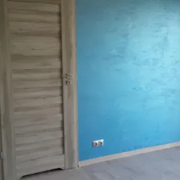 Ściana pomalowana na błękitno z efektem dekoracyjnym w Radomiu, widoczna drewnopodobna ościeżnica drzwiowa i szara podłoga.