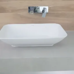 Biała, prostokątna umywalka nablatowa marki Vitra, zamontowana na szarym blacie z płytek, z chromowaną baterią i drewnopodobnymi płytkami na ścianie, widoczne puszki elektryczne w ścianie.