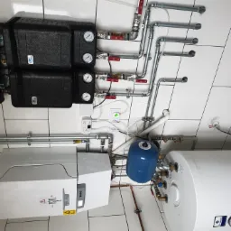 Kompleksowa instalacja kotła gazowego z rozdzielaczami, zasobnikiem CWU i naczyniem przeponowym, widoczne rury, zawory i manometry, białe płytki na ścianie.
