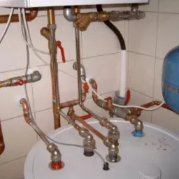 Kompletna instalacja wodna z miedzianymi rurami, zaworami i zbiornikiem ciśnieniowym w pomieszczeniu z wyłożonymi kafelkami ścianami. Widoczne złącza i elementy montażowe.