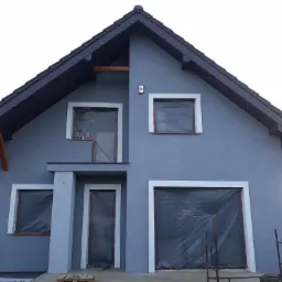 Dom jednorodzinny po nałożeniu szarego tynku, z oknami i drzwiami zabezpieczonymi folią, widoczny fragment rusztowania i paleta.