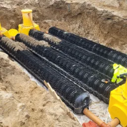 Instalacja drenażowa: cztery rzędy czarnych, tunelowych modułów drenażowych ułożonych w wykopie na białej geowłókninie, z żółtymi studzienkami kontrolnymi i rurami odprowadzającymi, na tle ziemi...