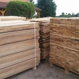 Ułożone na paletach i związane taśmą pakiety świeżo przetartego drewna w naturalnym, jasnym odcieniu, z widocznymi słojami i strukturą drewna. W tle zieleń drzew i krzew&oacute;w oraz fragment budynku...