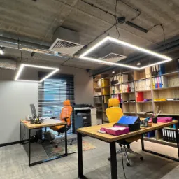 Nowoczesne biuro z kwadratowymi lampami LED pod sufitem, biurkami z drewnianymi blatami i kolorowymi krzesłami, widok na regał z segregatorami i drukarkę