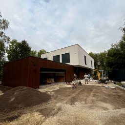 LEDBEST Sp. z o.o. - Nowoczesny dom z garażem w trakcie budowy. Widoczny mini-koparka, robotnicy i przygotowany teren. Ujęcie z dołu, pochmurne niebo w tle.