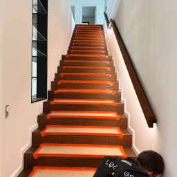 LEDBEST Sp. z o.o. - Montaż oświetlenia schodowego: pracownik instaluje listwy LED na schodach zabezpieczonych taśmą malarską. Widoczne podświetlone schody i fragment pracownika.