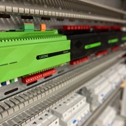 LEDBEST Sp. z o.o. - Panel sterowania inteligentnego domu Loxone z zielonym modułem głównym, widoczne bezpieczniki i listwy zaciskowe. Ujęcie z perspektywy, lekko rozmyte tło.