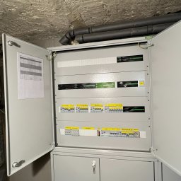 LEDBEST Sp. z o.o. - Otwarta szafa rozdzielcza z bezpiecznikami i dokumentacją, widoczne przewody i elementy instalacji elektrycznej. Szara obudowa, surowy strop w tle.