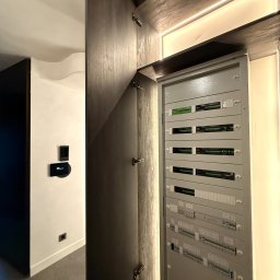 LEDBEST Sp. z o.o. - Otwarta szafa z szarą rozdzielnicą elektryczną w nowoczesnym wnętrzu. Widoczny panel sterowania inteligentnego domu na ścianie i oświetlenie LED w szafie.