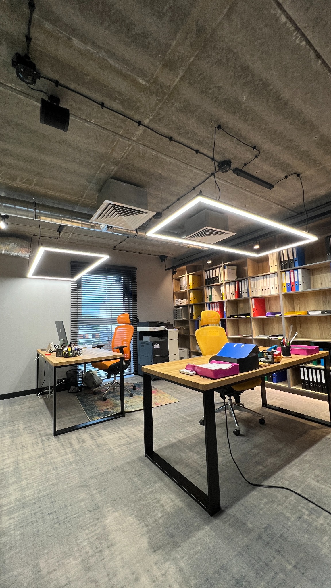 Nowoczesne biuro z kwadratowymi lampami LED pod sufitem, biurkami z drewnianymi blatami i kolorowymi krzesłami, widok na regał z segregatorami i drukarkę