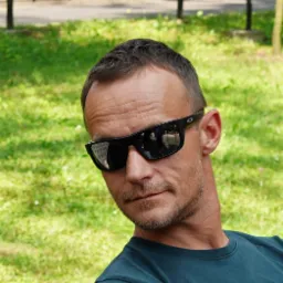 Mężczyzna w okularach przeciwsłonecznych, siedzący na tle zielonej trawy. W odbiciu okularów widoczny park, co sugeruje dbałość o otoczenie.