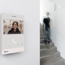 Installservice - Biały wideofon Golmar na ścianie, wyświetlający uśmiechniętą kobietę, z rozmytą postacią schodzącą po schodach w tle.