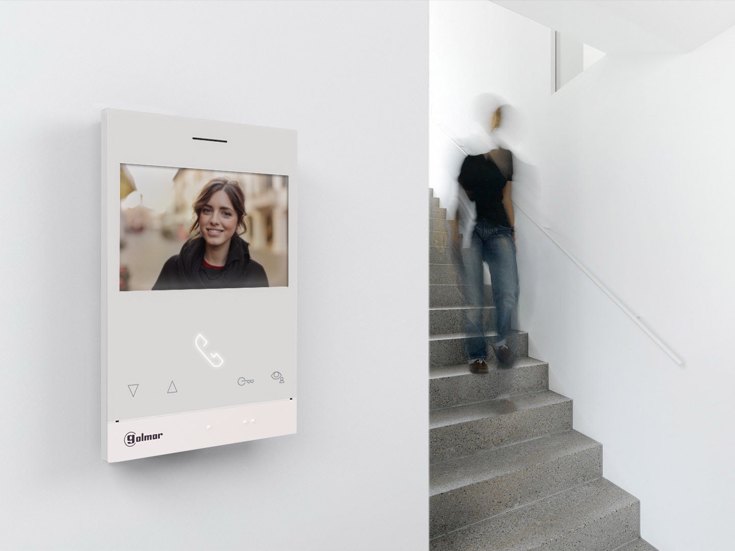 Biały wideofon Golmar na ścianie, wyświetlający uśmiechniętą kobietę, z rozmytą postacią schodzącą po schodach w tle.