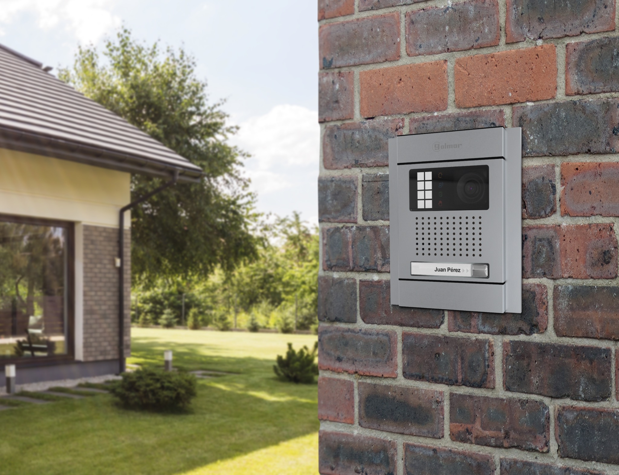 Szary wideodomofon Golmar z kamerą i klawiaturą numeryczną zamontowany na ceglanej ścianie domu z zadbanym trawnikiem w tle.