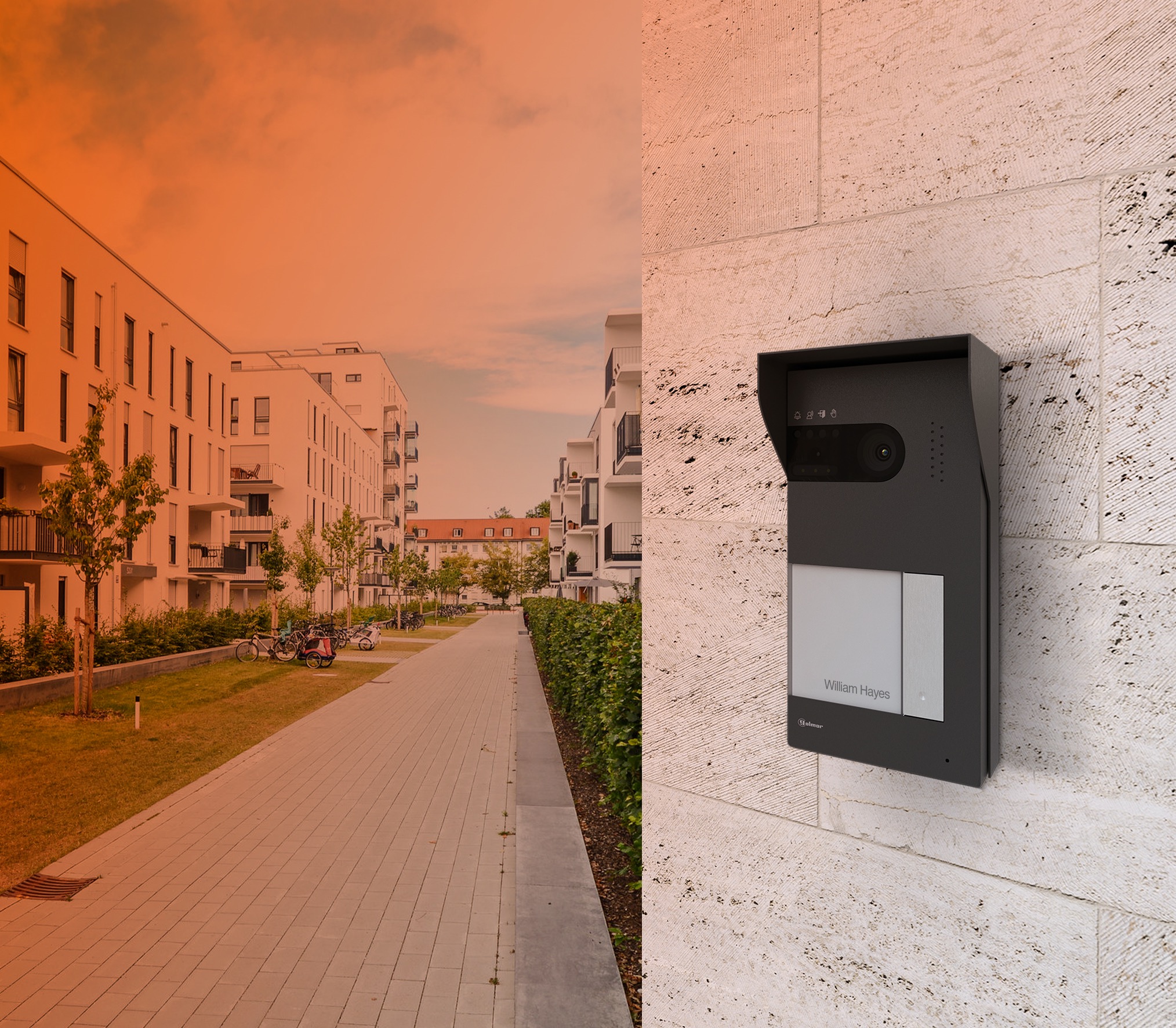 Nowoczesny wideofon w obudowie z daszkiem, zamontowany na elewacji budynku mieszkalnego z jasnego kamienia. W tle widoczny chodnik, zieleń i budynki mieszkalne.