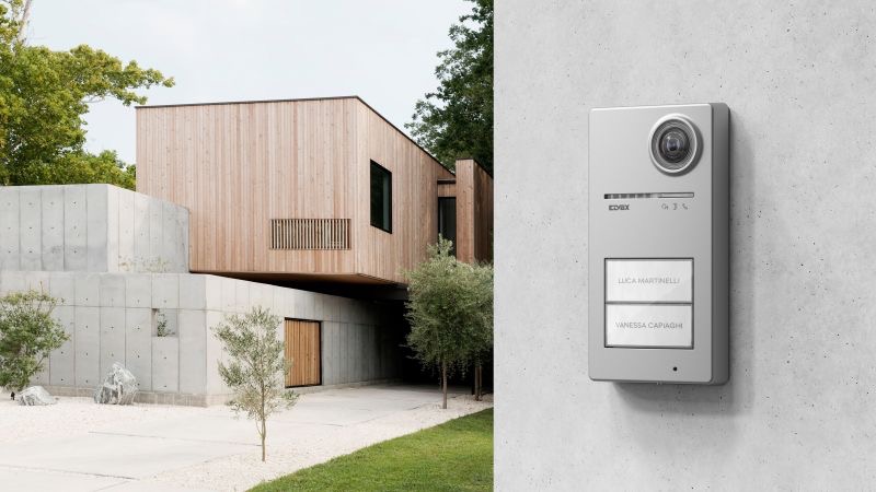Nowoczesny, srebrny wideodomofon z kamerą, zamontowany na betonowej ścianie na tle minimalistycznego domu z elementami drewna i betonu architektonicznego.