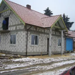 Dom w trakcie budowy, widoczne białe bloczki, czerwona dachówka, okna i pracownik na balkonie. Wokół śnieg i błoto. Częściowo ukończony budynek.