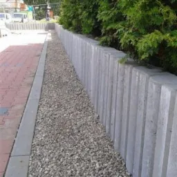 Szara palisada oddzielająca chodnik z czerwonej kostki od nasadzeń z krzewów iglastych, wypełniona drobnym żwirem.