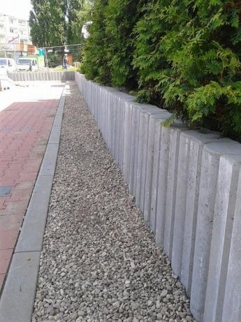 Szara palisada oddzielająca chodnik z czerwonej kostki od nasadzeń z krzewów iglastych, wypełniona drobnym żwirem.