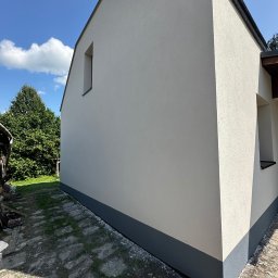 Ściana domu po renowacji elewacji, z widocznym fragmentem podmurówki w kolorze grafitowym. Jasna elewacja w kontraście z błękitnym niebem i zielenią drzew.