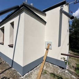 Jasna elewacja budynku z szarym cokołem i orynnowaniem, widoczna skrzynka na ścianie i oparta o nią deska. Błękitne niebo w tle.