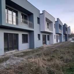 Rząd nowo wybudowanych, pomalowanych na różne odcienie szarości domów szeregowych z balkonami i dużymi oknami, widzianych z perspektywy trawnika.