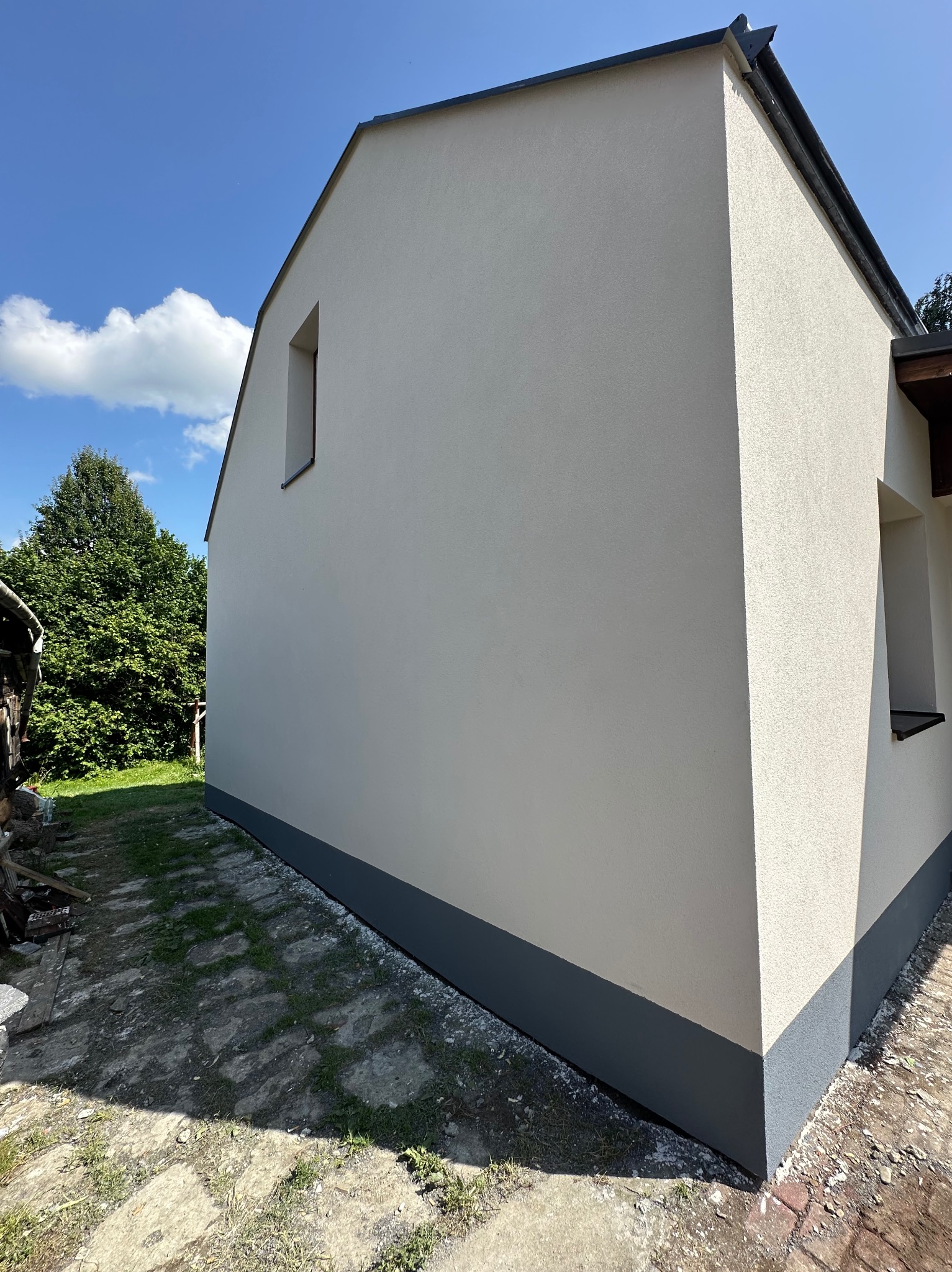 Ściana domu po renowacji elewacji, z widocznym fragmentem podmurówki w kolorze grafitowym. Jasna elewacja w kontraście z błękitnym niebem i zielenią drzew.