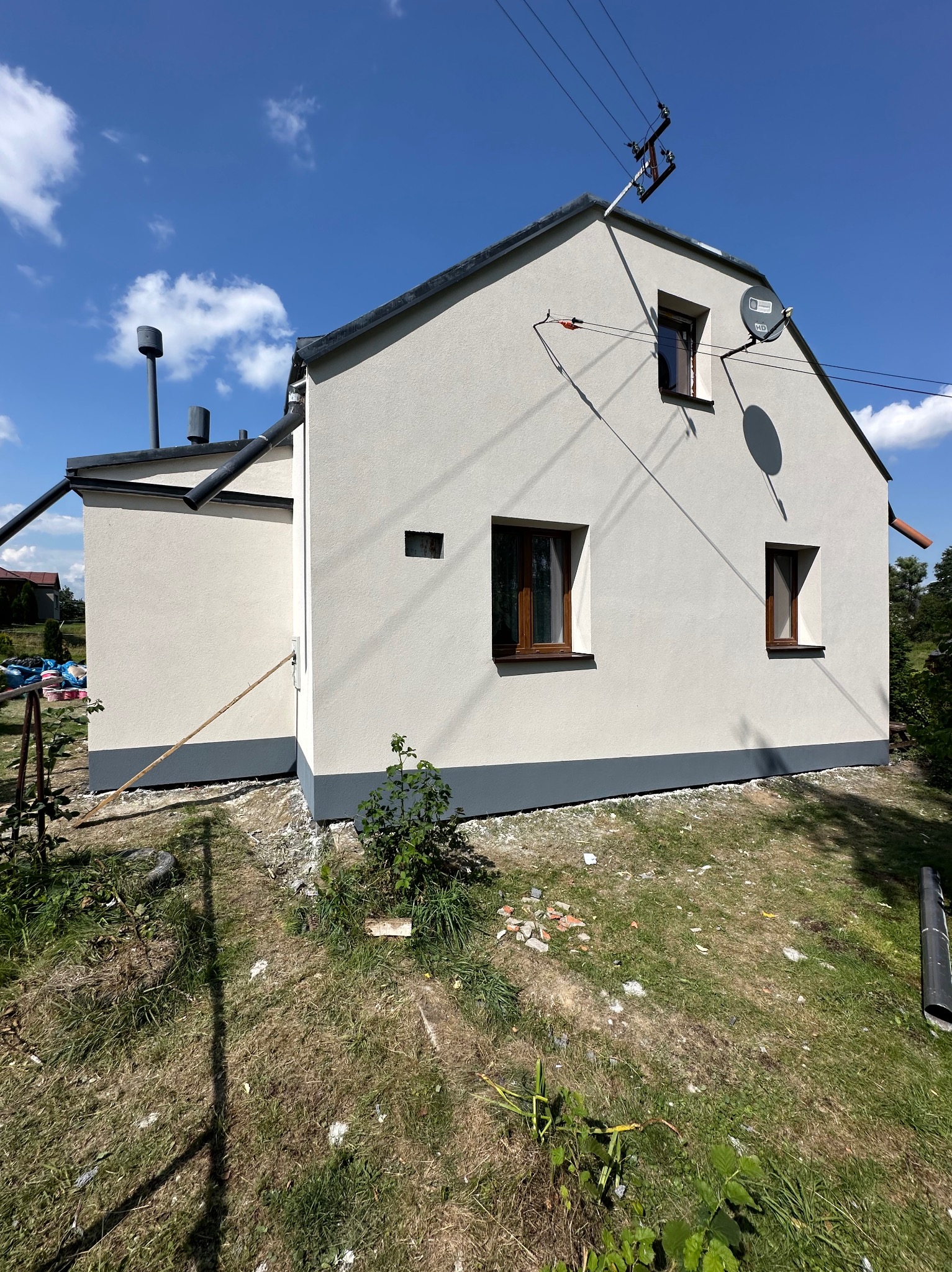 Jasny dom jednorodzinny po renowacji elewacji, z widoczną szarą podmurówką i nowymi oknami. Na dachu antena satelitarna. Błękitne niebo z chmurami w tle.