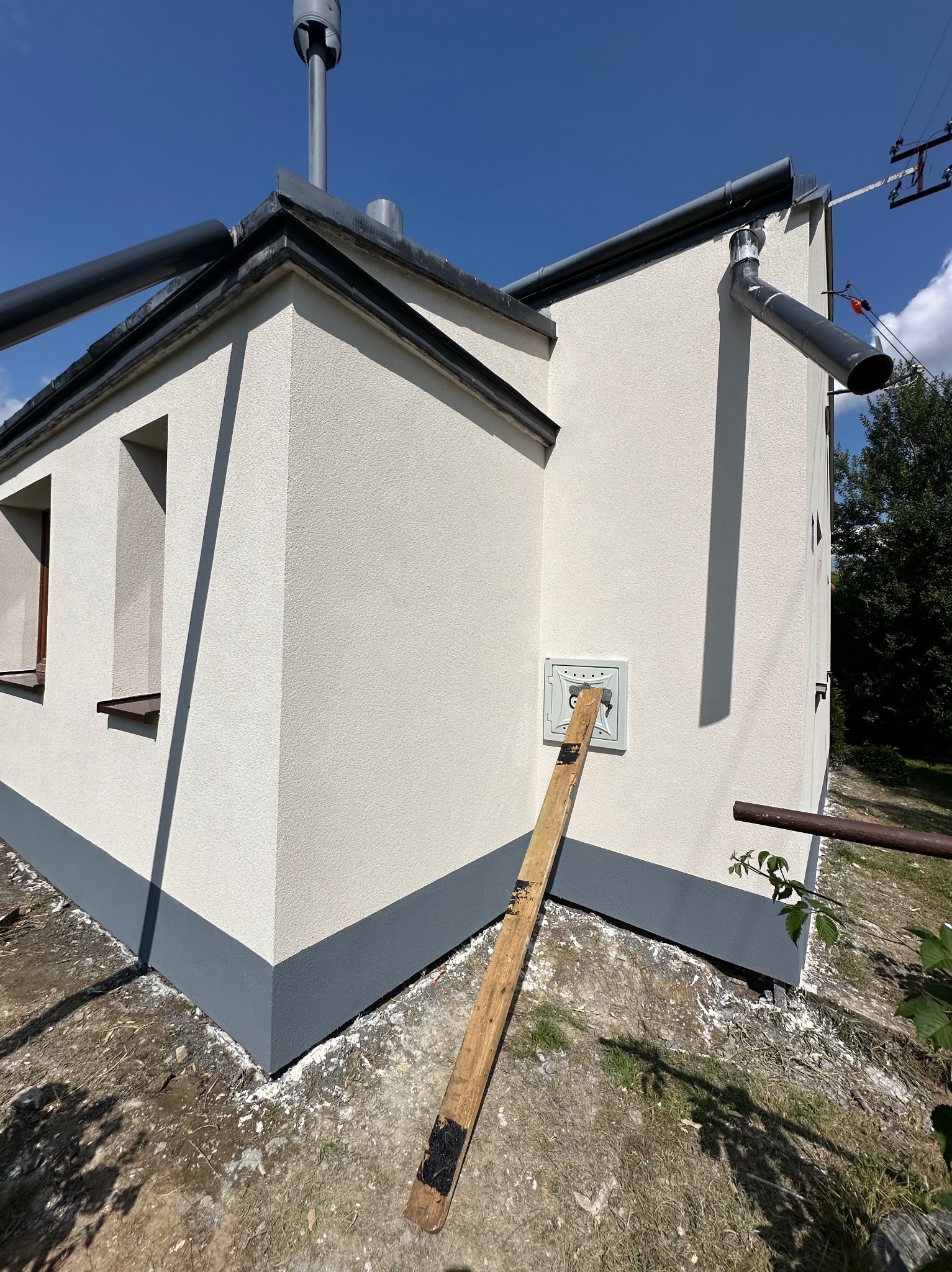 Jasna elewacja budynku z szarym cokołem i orynnowaniem, widoczna skrzynka na ścianie i oparta o nią deska. Błękitne niebo w tle.