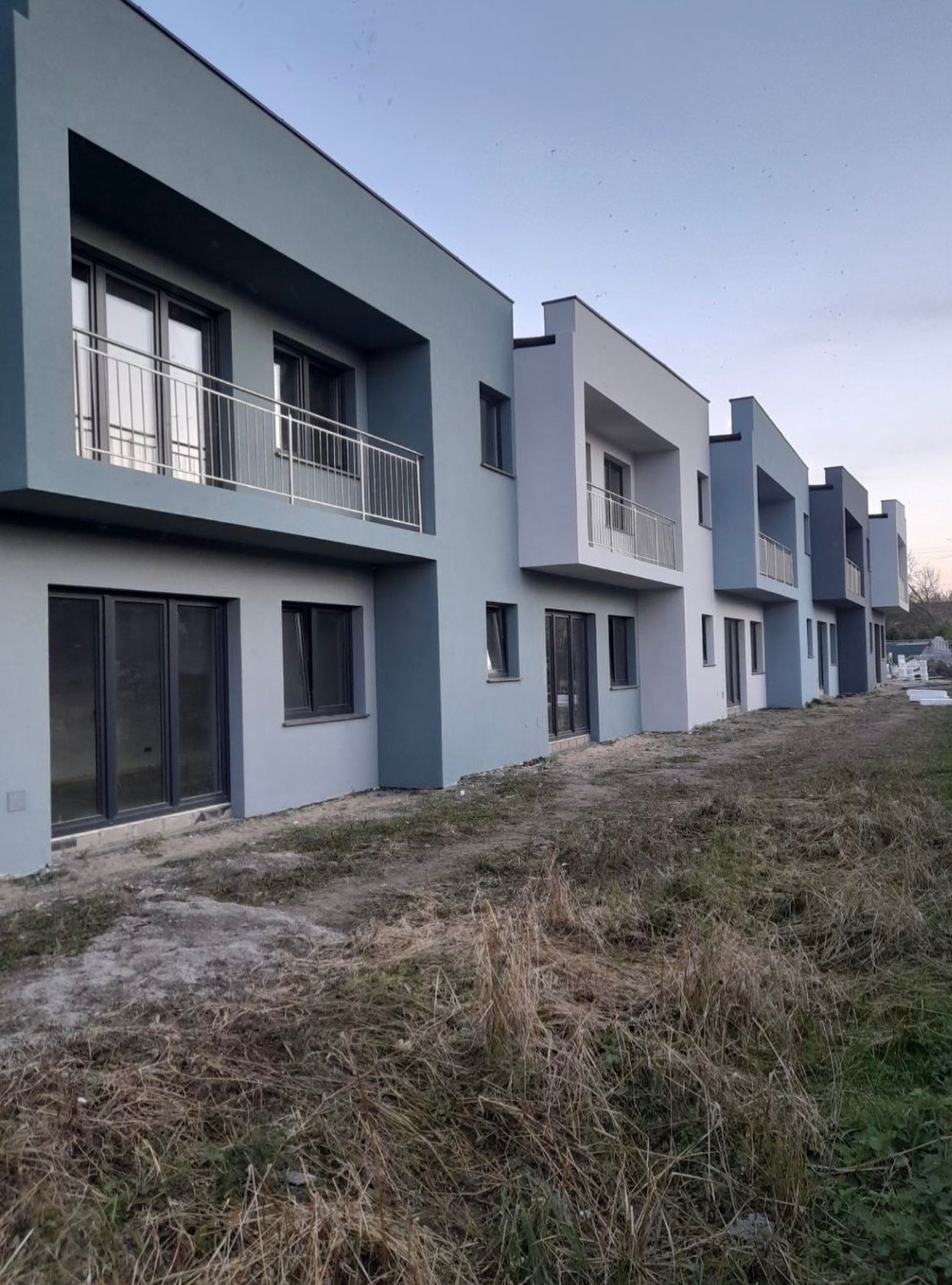 Rząd nowo wybudowanych, pomalowanych na różne odcienie szarości domów szeregowych z balkonami i dużymi oknami, widzianych z perspektywy trawnika.