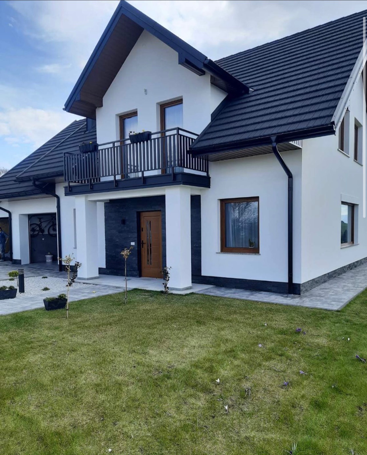 Nowoczesny dom jednorodzinny z balkonem i ciemną dachówką, biała elewacja z elementami grafitu, zadbany trawnik, błękitne niebo w tle.