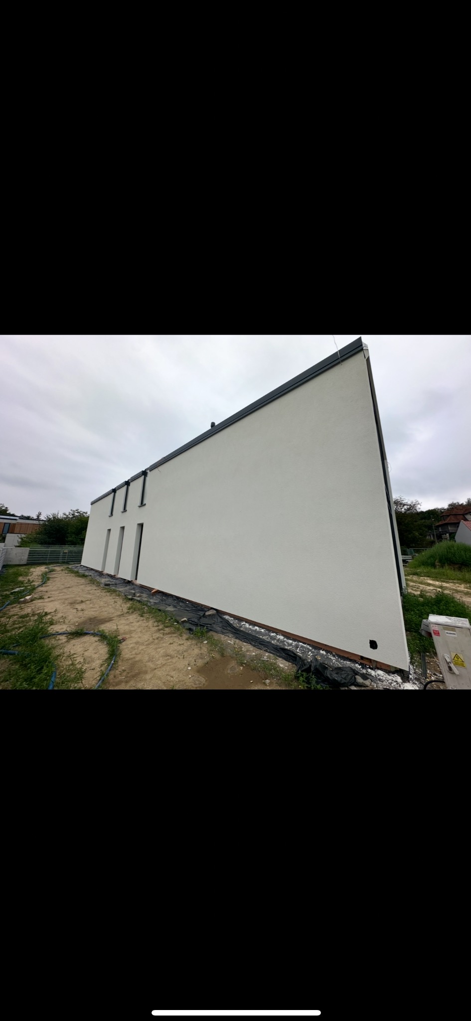 Biała, gładka elewacja domu jednorodzinnego z trzema oknami, widok z dołu, pochmurne niebo. Wykończona elewacja, teren wokół domu przygotowany pod zagospodarowanie.