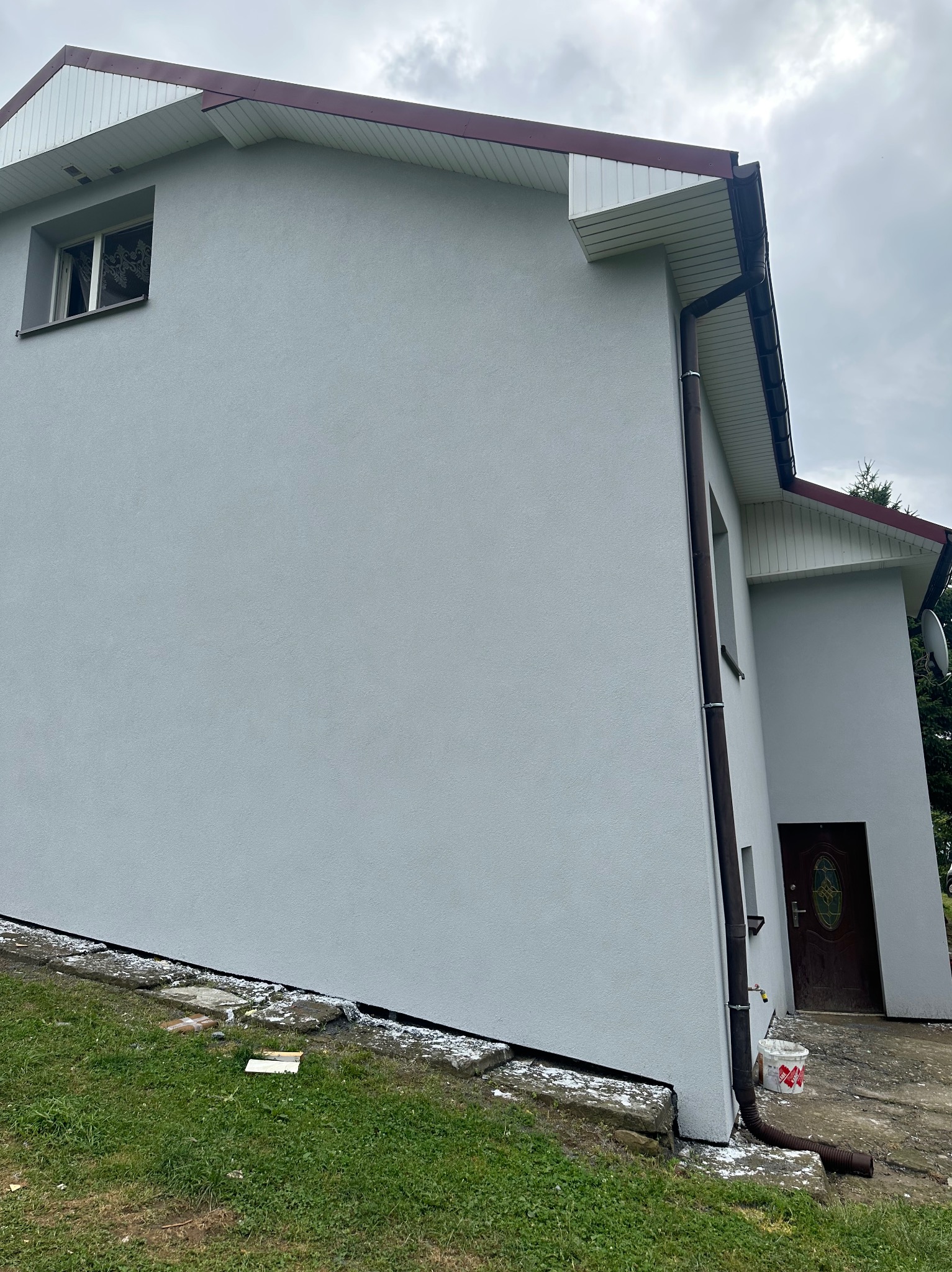 Jasnoszara elewacja domu jednorodzinnego z brązowymi rynnami i dachem. Widoczny fragment trawnika i wejście do budynku. Świeżo położony tynk.