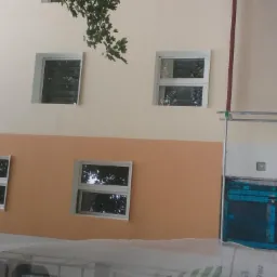 Elewacja budynku w trakcie odnawiania, widoczne rusztowania, okna z białymi ramami, fragmenty drzew i materiały budowlane w zielonych opakowaniach.