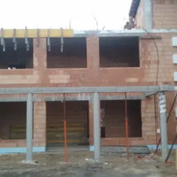 Surowy stan dwukondygnacyjnego budynku z czerwonej cegły, widoczne otwory okienne i drzwiowe, częściowo ocieplone ściany, szalunki na stropie i rusztowania z boku.