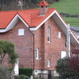 Dom jednorodzinny z czerwonym dachem ceramicznym, ceglaną elewacją i małym balkonem, otoczony zielenią i drzewami.