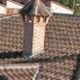 Brązowa dachówka ceramiczna ułożona na dachu z widocznym kominem z cegły, oświetlona naturalnym światłem.