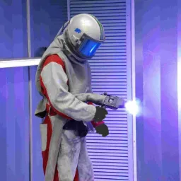 Pracownik w kombinezonie ochronnym i hełmie, trzymający pistolet do czyszczenia strumieniowego w jasnym, przemysłowym otoczeniu z niebieskimi panelami.