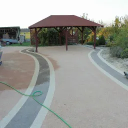 Świeżo ułożona nawierzchnia z żywicy epoksydowej w ogrodzie z altaną, widoczne narzędzia i wózek budowlany.