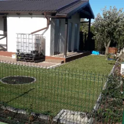 Nowoczesny ogród z zielonym trawnikiem, zbiornikiem na deszczówkę przy budynku, drewnianym ogrodzeniem i zieloną siatką ogrodzeniową.