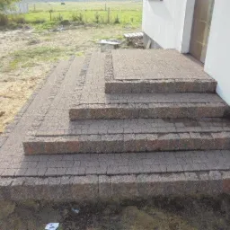 Schody wejściowe do domu wyłożone kostką granitową w odcieniach brązu, otoczone ziemią i trawą, z widocznym fragmentem elewacji budynku.