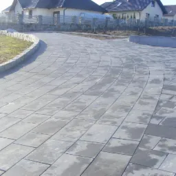 Szeroka, wyłożona szarą kostką brukową droga dojazdowa do domów jednorodzinnych, z kamiennym obrzeżem i elementami małej architektury krajobrazu w tle.