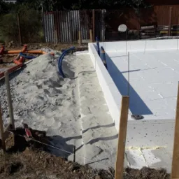 Przygotowanie terenu pod fundament w Głogowie: białe płyty izolacyjne ułożone w wykopie, zasypane piaskiem, z widocznymi rurami i drewnianym szalunkiem.