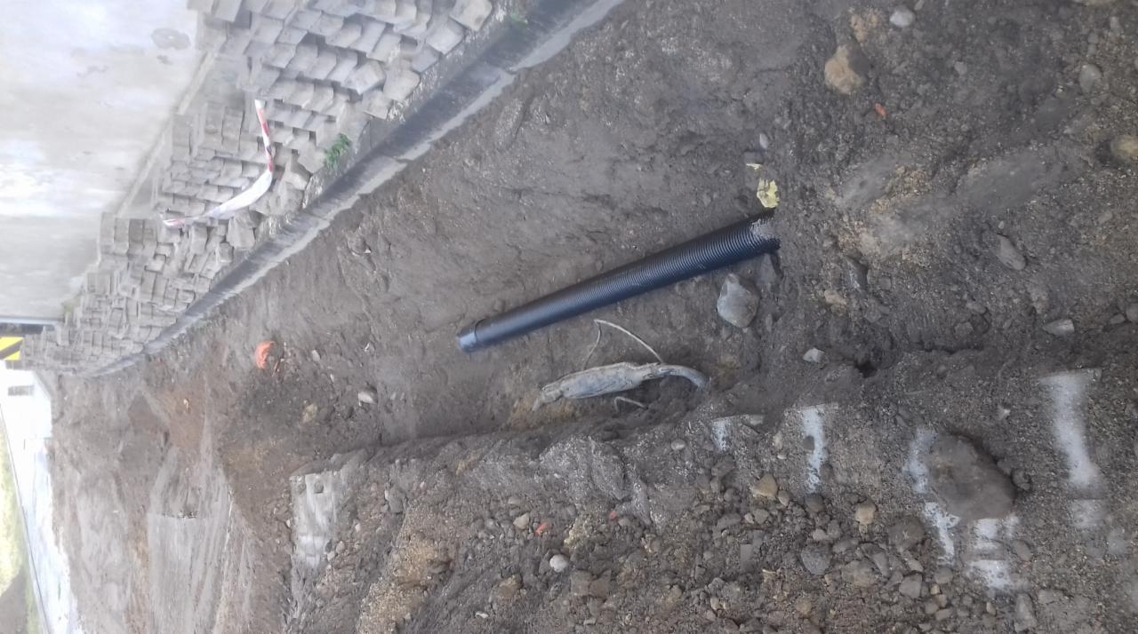 Wykop w ziemi z widoczną czarną rurą karbowaną, fragmentem krawężnika z kostki i białymi oznaczeniami na gruncie.