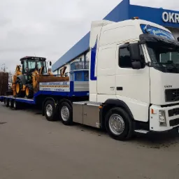 Biała ciężarówka Volvo z niskopodwoziową naczepą przewozi żółto-czarną koparko-ładowarkę na tle budynku z napisem 'OKRĘGOWA'.