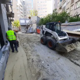 Operator w kamizelce odblaskowej obsługuje zagęszczarkę obok fundamentów budynku; w tle widoczny mały, biały Bobcat i budynki mieszkalne.