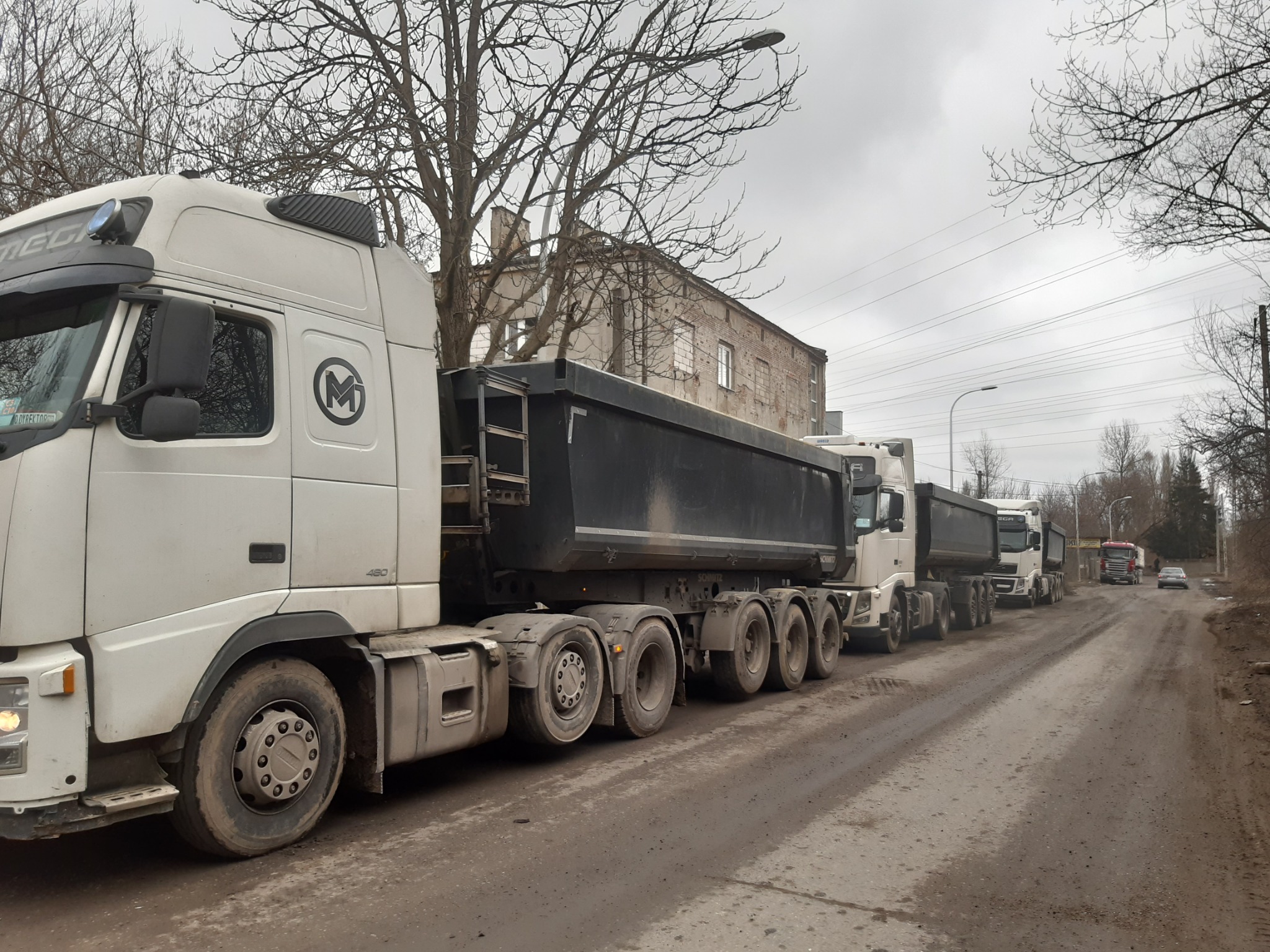 Szereg białych ciężarówek z czarnymi naczepami typu wywrotka stoi na błotnistej drodze w pochmurny dzień, widoczny budynek w tle i drzewa bez liści.