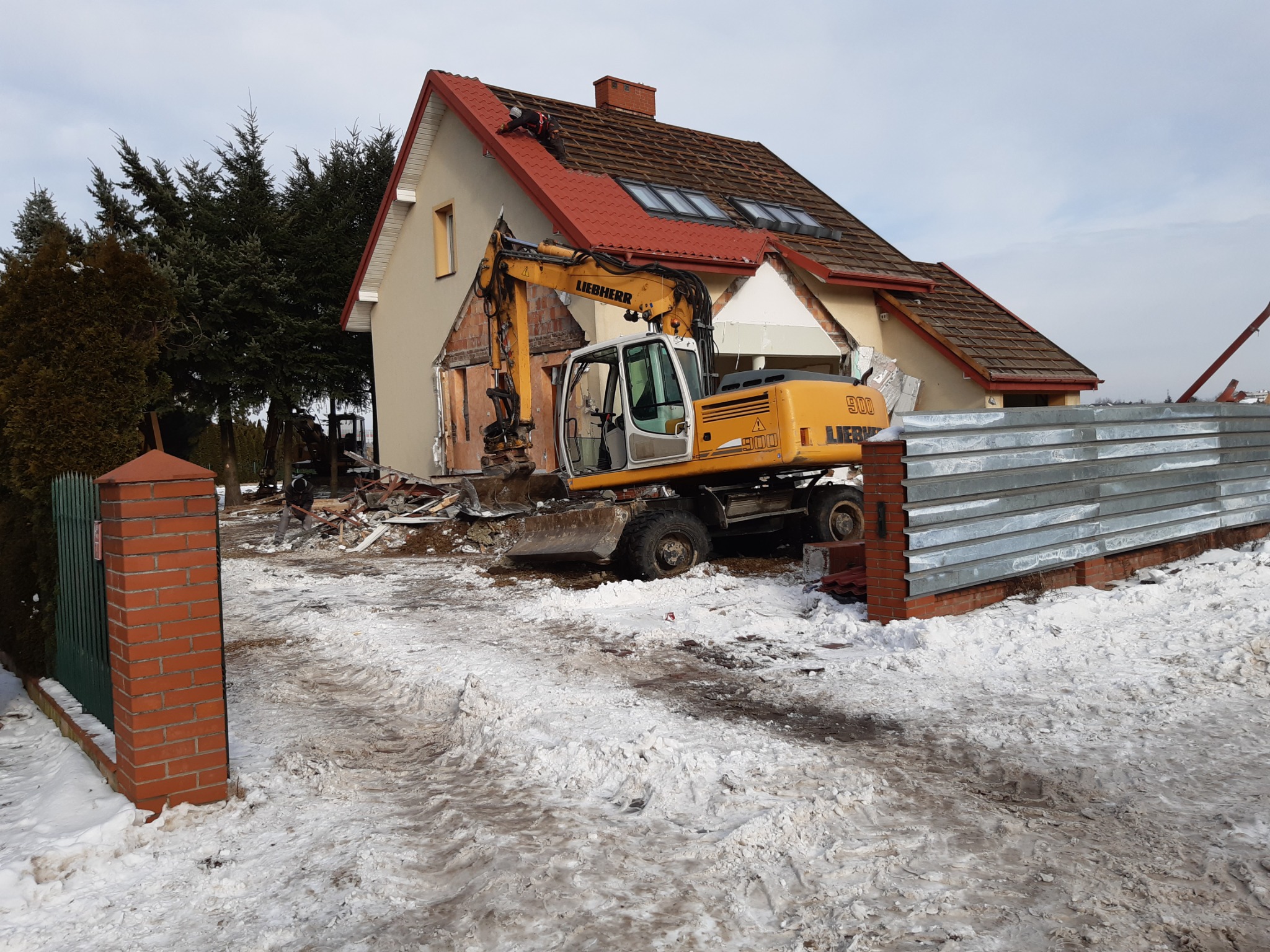 Żółta koparka Liebherr 900 częściowo rozbiera murowany dom z czerwoną dachówką, fragmenty ścian i dachu leżą na ośnieżonym terenie.