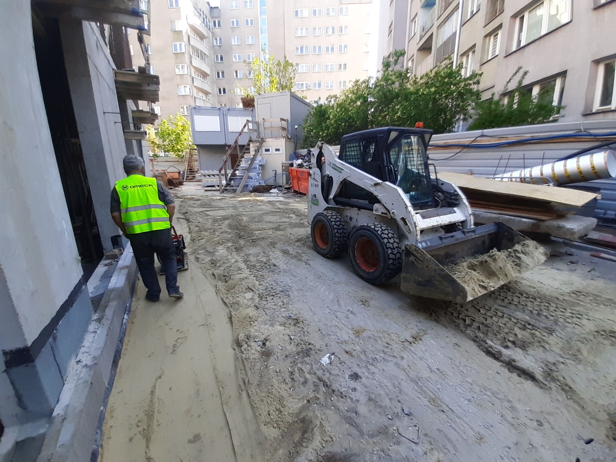 Operator w kamizelce odblaskowej obsługuje zagęszczarkę obok fundamentów budynku; w tle widoczny mały, biały Bobcat i budynki mieszkalne.